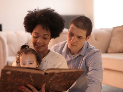 A man and a woman sit on the floor in front of a couch. They are reading to a toddler sitting in the woman's lap.