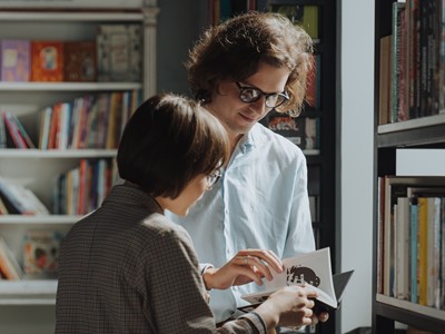 A photo of two people standing in a library, they are looking at a passage out of a book together 