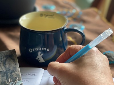 A photo of someone holding a pen and writing, there's a second book and mug with a warm drink.