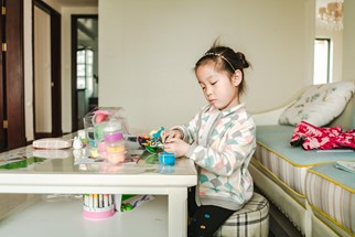 A preschool aged child sits in a loungeroom with various craft supplies in front of her on a table. She is looking down absorbed in making a colourful creation